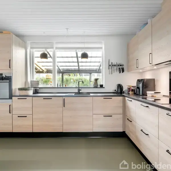 Køkken med lyse træskabe; opvaskemaskine, kaffemaskine, og køkkenredskaber på sorte bordplader; stort vindue med udsigt til en overdækket terrasse og grønne omgivelser.