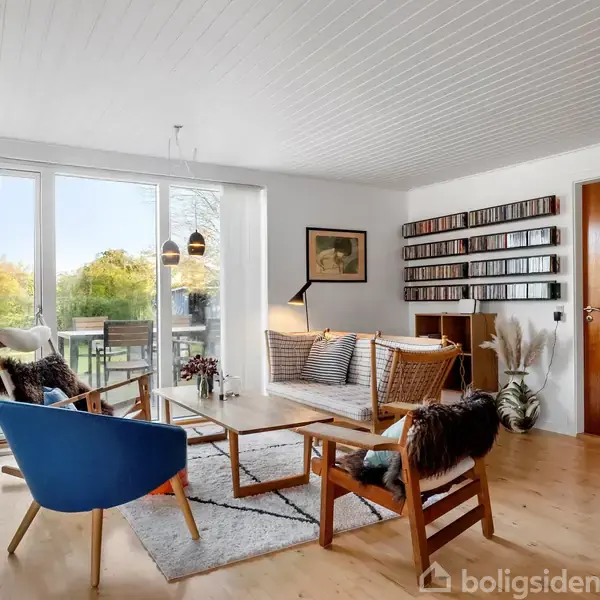 En stue med træmøbler og en blå stol rundt om et sofabord. Store vinduer viser en terrasse og have udenfor. Væggene er dekoreret med billeder og en cd-reol.