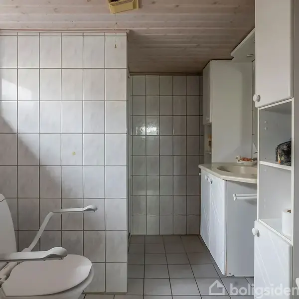 Toilet med håndtag står i et flisebelagt badeværelse. Til højre ses en vask med skab og hylder. Rummet er lyst op af et lille ovenlys vindue.