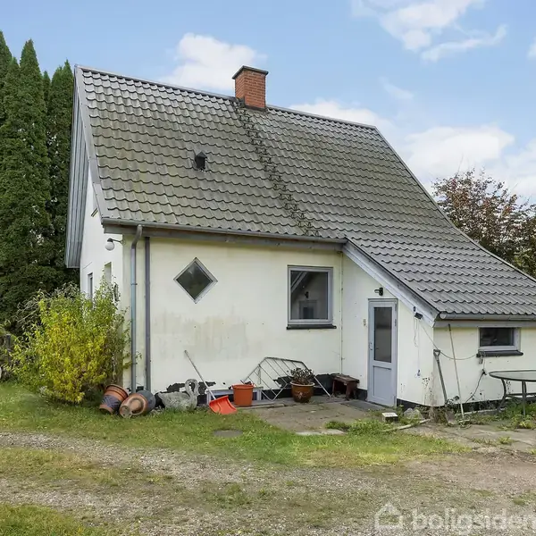 Et lille hus med gråt tag og hvid facade står stille i en grøn have omgivet af buske og træer. En grussti leder op til hoveddøren.