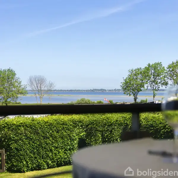 En flaske og to vinglas står på et bord, med fokus mod en udsigt over en sø omgivet af grønne træer under en klar blå himmel.