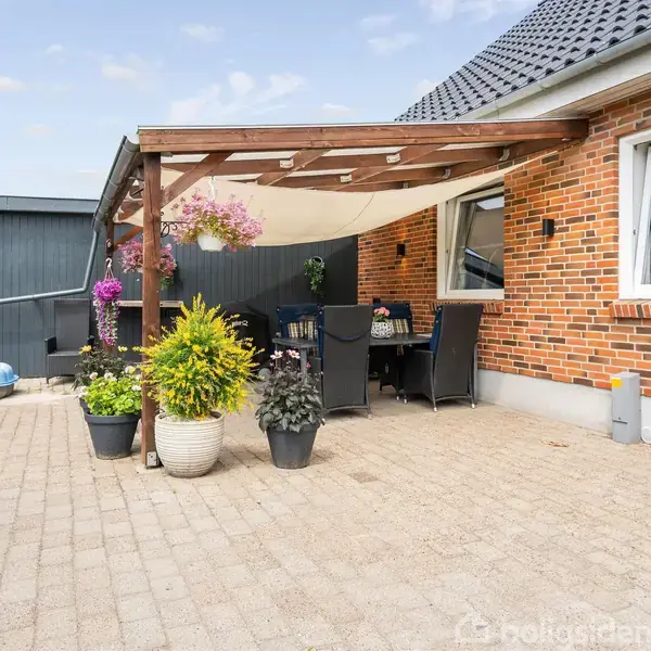 En terrasse med borde og stole står under en pergola i træ, omgivet af blomsterkrukker, op ad en rødstensbygning. Skaber et hyggeligt udendørs opholdsområde ved huset.