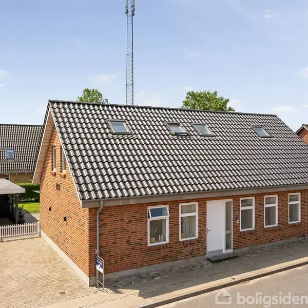 En rød murstenshuse står på en brolagt gade, med et sort tegltag og flere vinduer. Nabohuse og en carport ses i baggrunden under en klar blå himmel.
