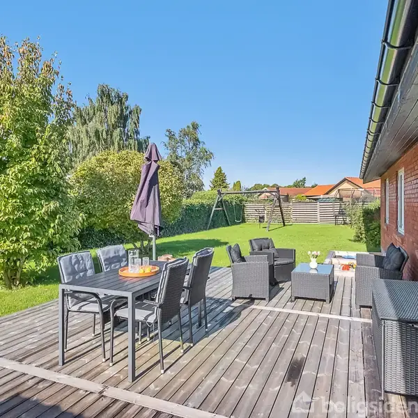 Terrasse med havemøbler står på en træplatform. Bord med stole har en parasol. Græsplæne omgivet af hække og træer i baggrunden. Husvæg til højre.