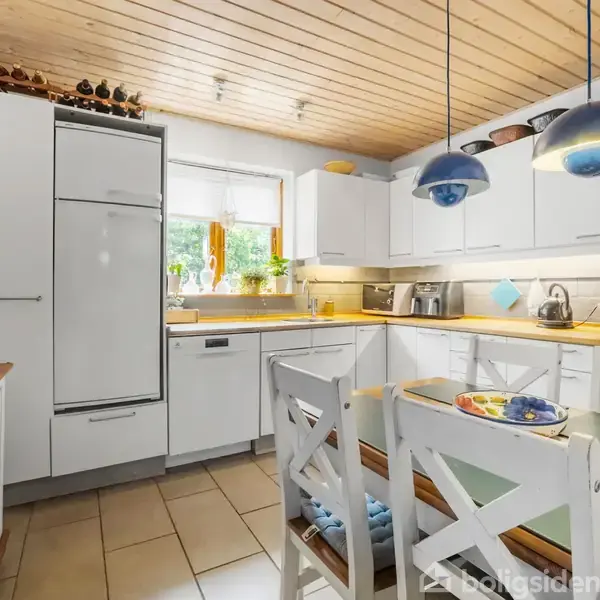 Køkken med hvide skabe og træbordplader. To blå lamper hænger over et spisebord med tallerkener. Rummet har vinduer, kakler på gulvet og en kaffemaskine på en bordplade.