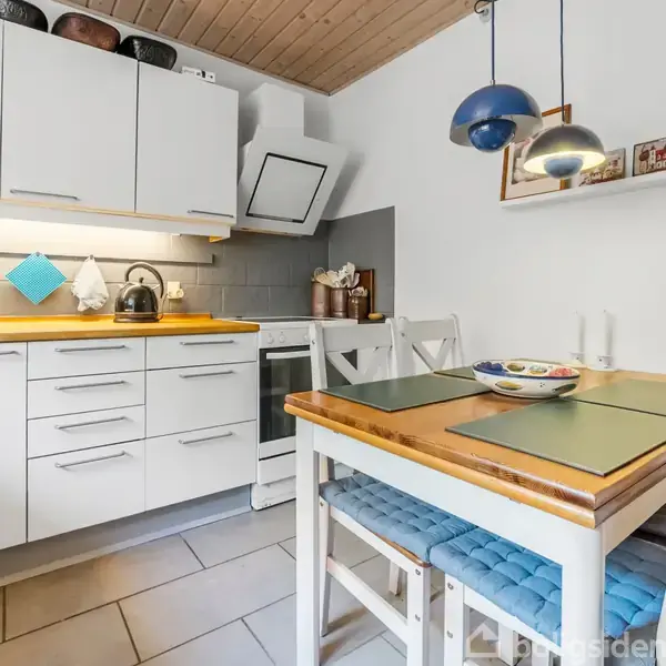 Et lyst køkken med hvide skabe og træbordplade. Et spisebord med dækkeservietter og en skål står ved siden af en blå polstret stol. Hyggebelysning fra lamper ovenover.