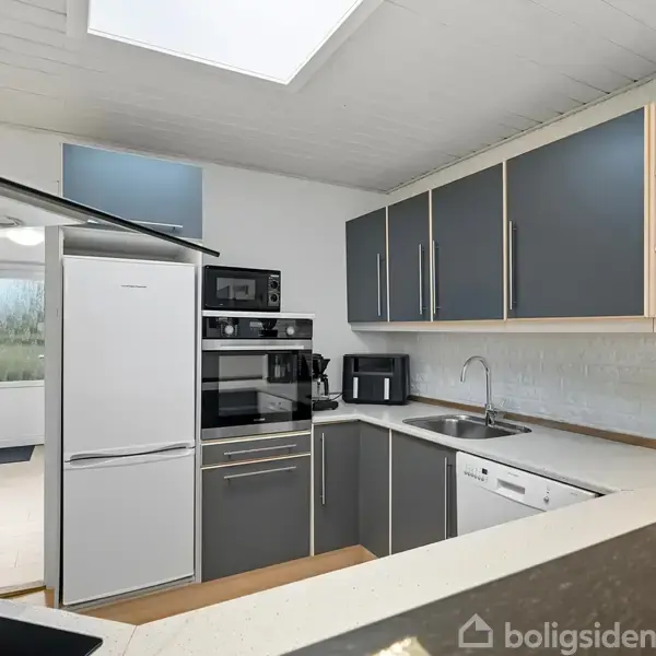 Et moderne køkken har grå skabe, en hvidkøleskab, en indbygget ovn, mikrobølgeovn og opvaskemaskine. Der er en sort kaffemaskine og planter ved vinduet. Skabe er delvist beklædt med træ.