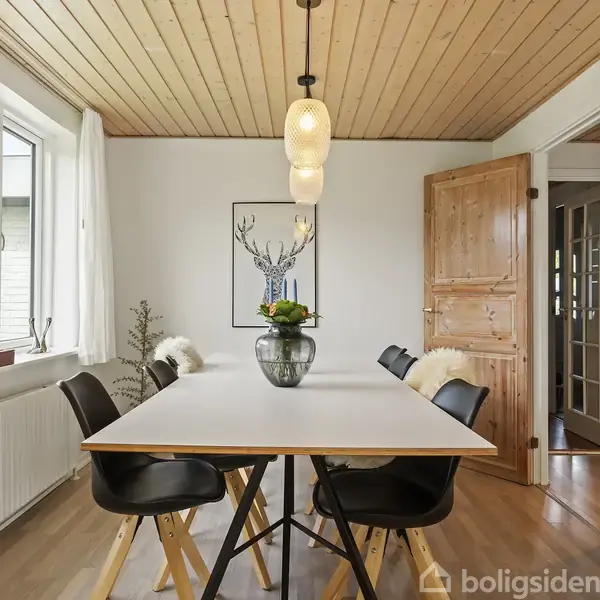 Spisebord med seks sorte stole omkring det, placeret i et lyst rum med trægulv og træbeklædt loft. Et vindue til venstre og en dør til højre danner konteksten. En stilfuld lampe hænger over bordet.
