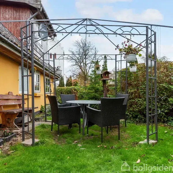 En metalpavillon står på en græsplæne med et bord og fire stole placeret under, omringet af grønne buske og et gulmalet hus til venstre. En bænk ses ved huset.