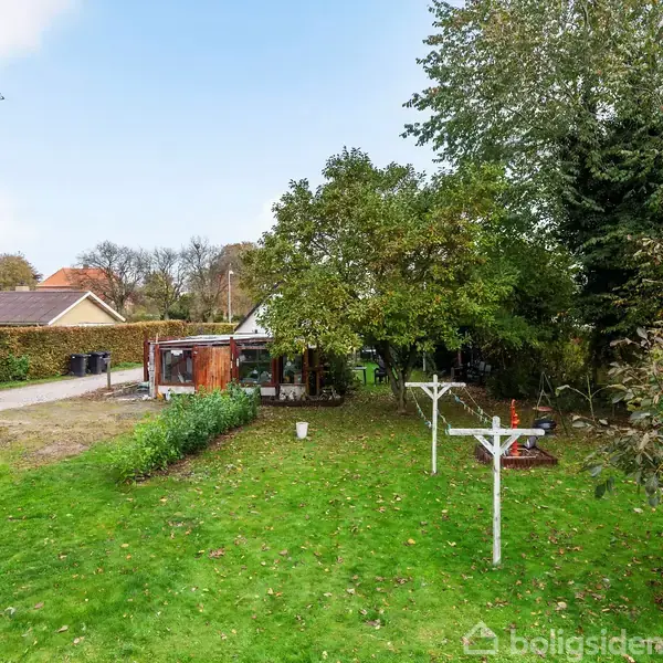 Græsplæne med to hvide tørrestativer står stille i en have omgivet af tætte træer. Små skure vises til venstre nær en lille vej i et villakvarter.