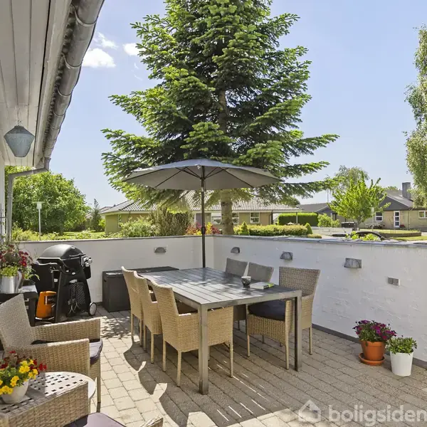 Udendørs terrasse med bord og stole under en parasol, omgivet af blomsterkrukker og placeret foran et hvidt hus. En grill står i hjørnet, og en stor træ ses i baggrunden.