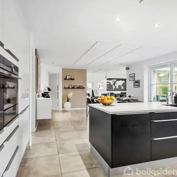 Et moderne køkken med en stor sort køkkenø fyldt med frugter. Køkkenet har indbyggede ovne til venstre og er lyst op af naturligt lys fra store vinduer mod højre.