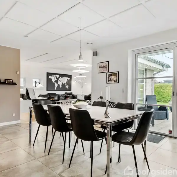 Et spisebord med sorte stole står på klinkegulv i en moderne spisestue med åbne hylder og dekorationer. To lamper hænger over bordet, og store vinduer fører til en terrasse.