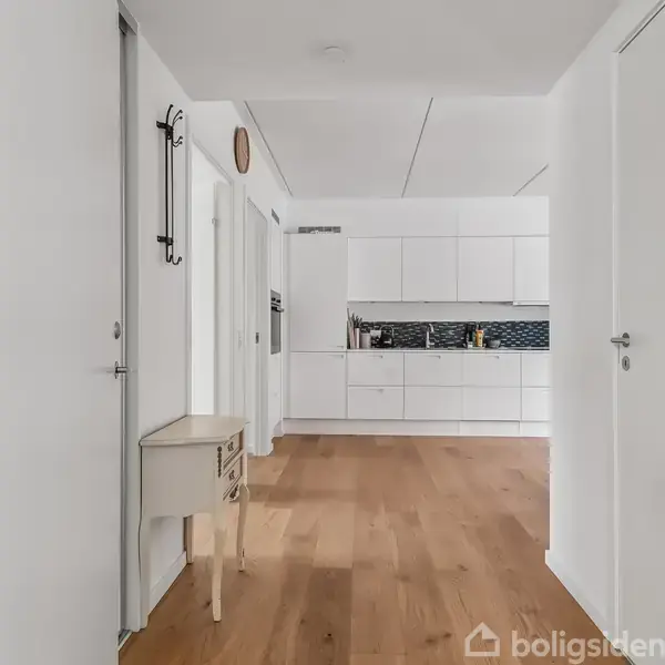 Køkken i minimalistisk hvidt design med træparketgulv og indbyggede skabe, set fra en gang med en lille konsolbord til venstre.