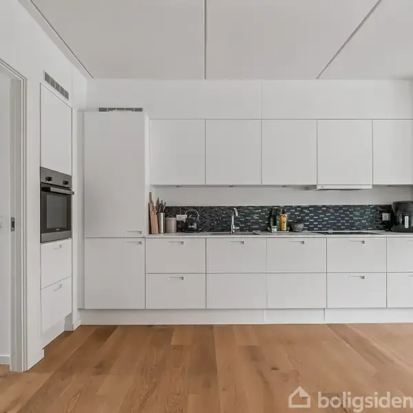 Et moderne køkken med hvide skabe og skuffer, indeholder ovn og små køkkenredskaber. Køkkenet har en mørk mosaik backsplash. Rummet har trægulv og en lys, enkel indretning.