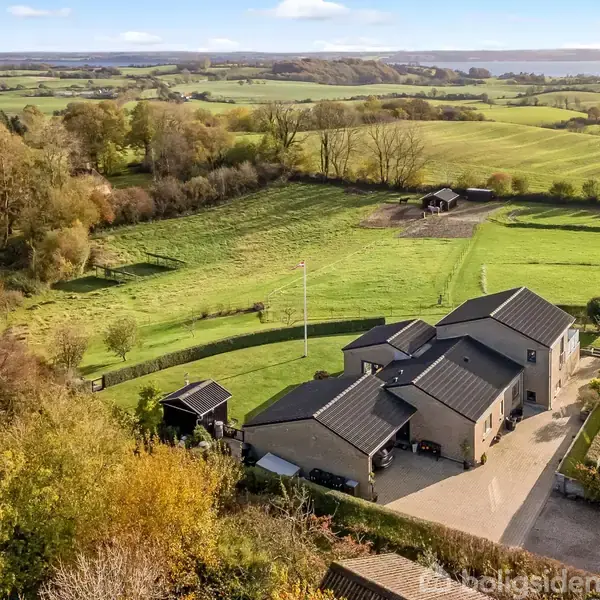 Et stort hus omgivet af grønne marker og træer ligger i et landligt område. Tæt ved huset er en grusvej og mindre bygninger, med synlige bakker i baggrunden.