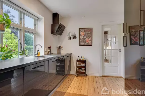 Køkken med sorte skabslåger og træbordplade. Knive hænger på væggen. Store vinduer til venstre giver udsigt til have. Plakater på væggen viser madmotiver.