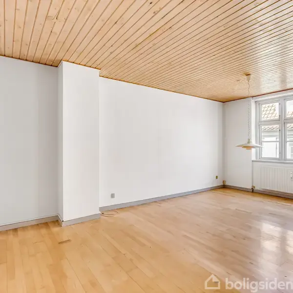 Et tomt rum med lyse trægulve og træloft. Rummet har hvide vægge og et vindue med flere ruder. Radiator under vinduet, minimal indretning, og hængelampe i hjørnet.