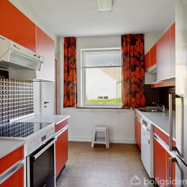 Køkken med orange skabe og komfur, handling i form af madlavning. Konteksten er en varm, retro indretning med blomstermønstrede gardiner og metalfliser bag komfuret. Ingen tekst til stede.