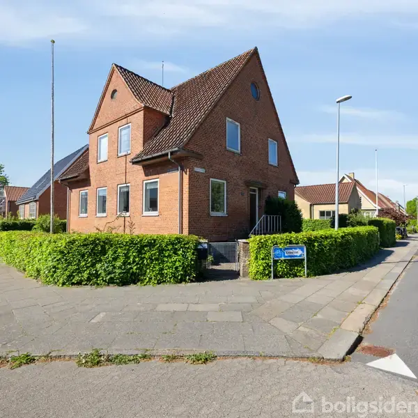Et rødt murstenshus står ved en vej omgivet af grønne buske. Huset har et skråt tag med solpaneler, og en blå ejendomsmægler-skilt står ved indkørslen på fortovet.