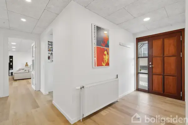 En lys gang med trægulv, en radiator under et kunstplakat med teksten "ANDY WARHOL LOUISIANA". Til højre ses en brun hoveddør. I baggrunden er der adgang til opholdsrum.