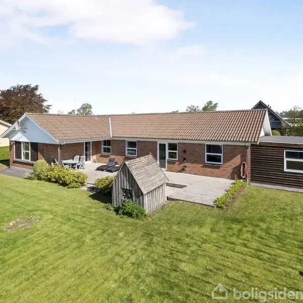 En rød murstensvilla med grå tagsten står stille. Husets baghave har en stor terrasse, grøn græsplæne og et lille skur. Omkringliggende huse og træer er synlige i baggrunden.