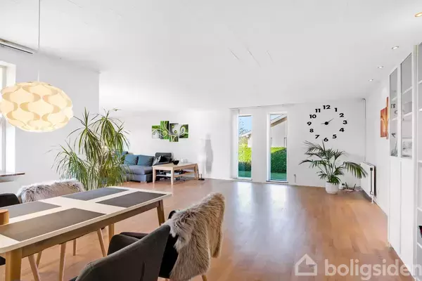 Stue med spisebord foran, dekoreret med pelsovertrukne stole, en stor pendellampe, planter og et ur på væggen. Baghaven ses igennem glasdørene i lyst rum.