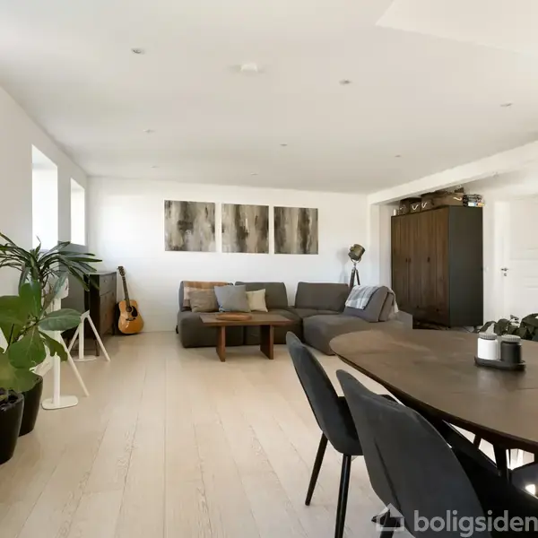 En lys stue med en grå sofa og et mørkt spisebord i forgrunden. Der er kunst på væggen, en guitar i hjørnet og planter som dekoration.