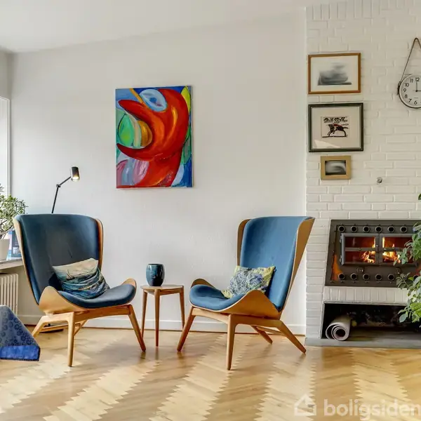 To lænestole vender mod hinanden ved et kaffebord i et stuehjørne. Der hænger farverigt kunst på væggen, og en hyggelig brændeovn står ved siden af vinduet med udsigt til en have.
