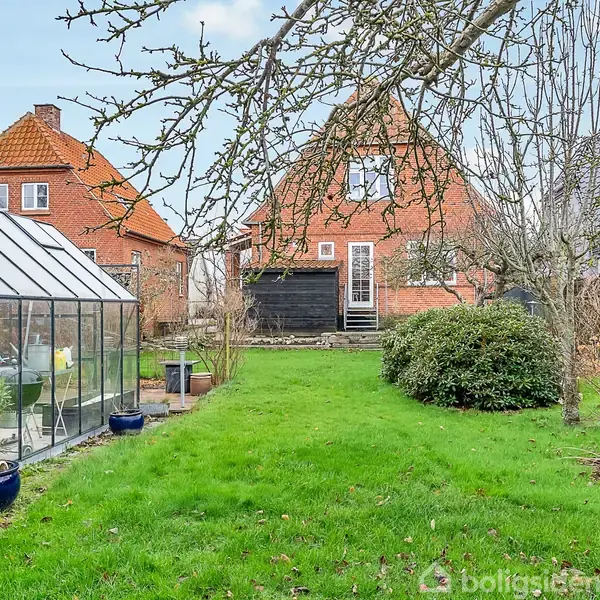 Rødt murstenshus med to etager og sorte skodder, stående i en have med grønt græs. Et drivhus er placeret til venstre, omgivet af buske og træer.