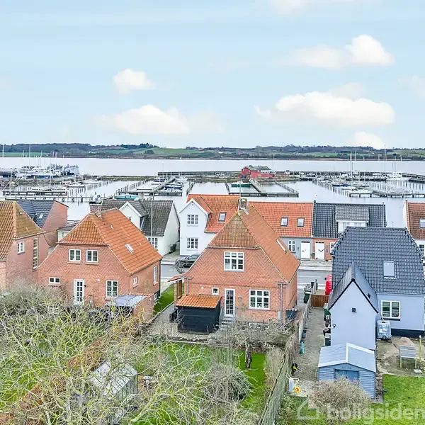 Rødstenshuse i forgrunden, tæt ved en marina med både til kaj. Vindstille himmel over en rolig vandflade. Husene har haver og er omgivet af stier og vegetation.