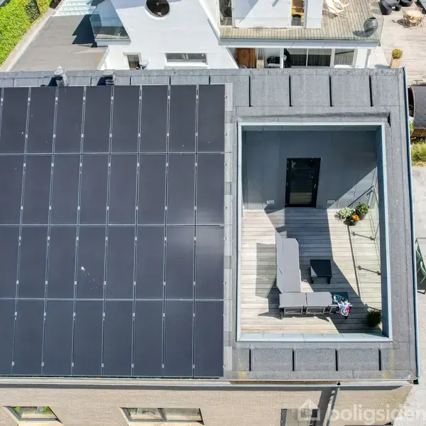 Solpaneler dækker taget af en moderne bygning med fladt tag. Ved siden af ligger en tagterrasse med liggestole og planter. Bygningen er omkranset af en grøn have.