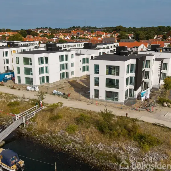 Flere moderne, hvide boligkomplekser står tæt ved en havn. Området omkring bygningerne synes under udvikling med nogle byggematerialer til stede. I baggrunden ses en by med røde tage.