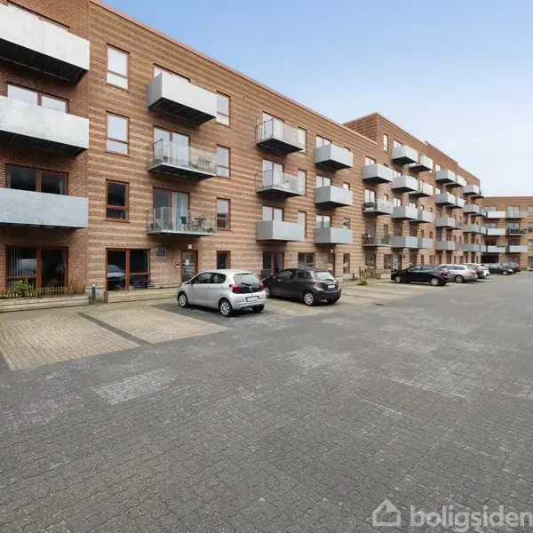 En række etagebygninger med altaner, står side om side. Foran bygningerne holder biler parkeret på en asfalteret parkeringsplads under en klar, blå himmel.