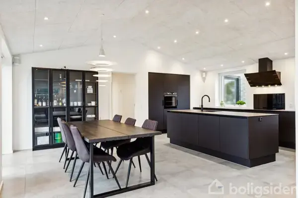 Et moderne køkken med sort spisebord og seks stole. Køkkenø med indbygget vask i en åben planløsning. Glasskab i baggrunden rummer flasker. Store vinduer giver naturligt lys.