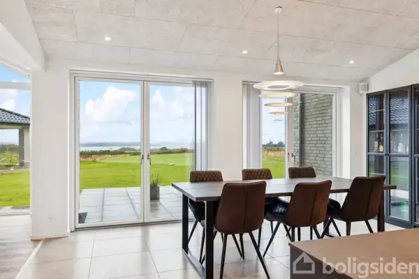 Spisebord med seks stole står i en lys spisestue. Rummet har store glasdøre, som åbner ud til en terrasse og grøn have med vid udsigt over landskabet. Pendellampe hænger over bordet.