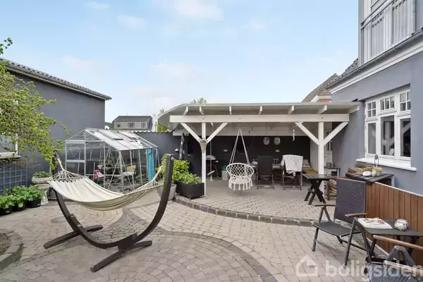 Hængekøje står i en flisebelagt gårdhave ved siden af et drivhus. En overdækket terrasse med stole og et bord skaber en hyggelig udendørs opholdsplads.