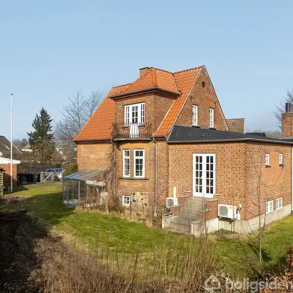 Et rødt murstenshus med et rødt tegltag står stille i en åben have. Omkring huset er der en kortklippet græsplæne og enkelte buske. En flagstang ses til venstre.
