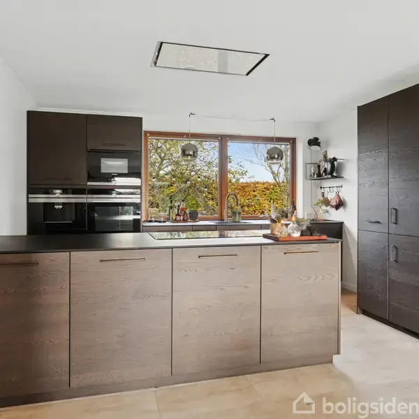 Moderne køkken med mørke træskabe, en indbygget ovn og kogeplade. Store vinduer med udsigt til træer tilfører naturligt lys. Væggene er hvide, og gulvet er flisebelagt.