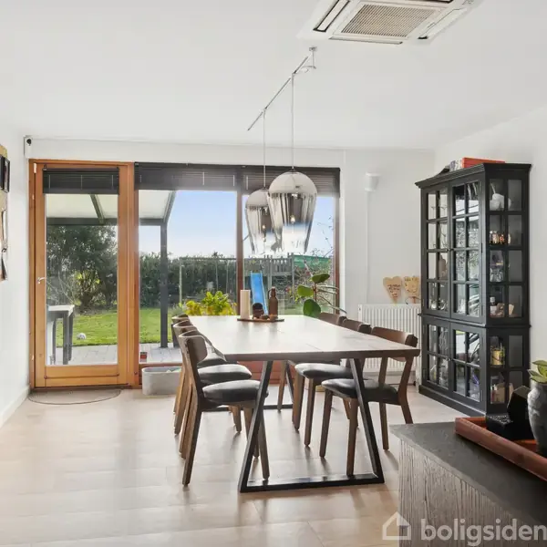 Spisebord med stole står i en lys spisestue med store vinduer, der åbner sig til en grøn have. Skab med glaslåger står ved siden af, og der hænger lamper over bordet.