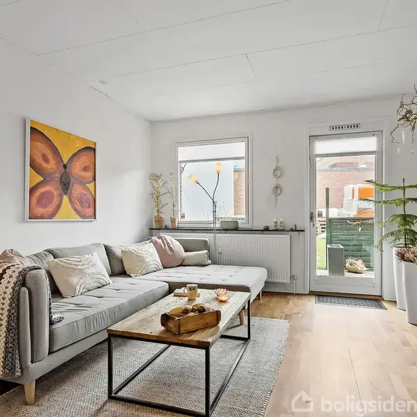 Grå sofa med puder og tæppe står i en lys stue. Et træbord med dekorationer er foran sofaen. Vægkunst hænger over sofaen, og vinduer giver udsigt til en have.