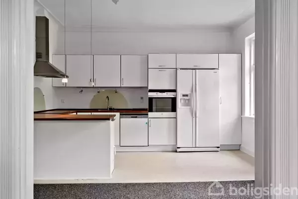 Et moderne køkken med hvide skabe, en stor køle-fryseskab og indbygget ovn. Køkkenbordet har en vask, og rummet er lyst med vinduer til højre.