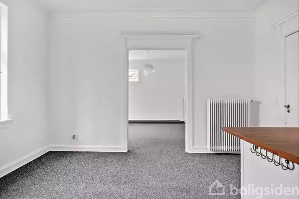 Et tomt rum med grå tæpper, en åben døråbning fører til et tilstødende værelse. En radiator er monteret på højre væg, og en træbordplade ses delvist.