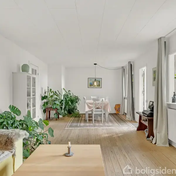 Stue med lyse træmøbler dominerer, med en sofa og sofabord i forgrunden. Store vinduer giver udsigt til en grøn baghave, omgivet af indendørs planter og spisestue.