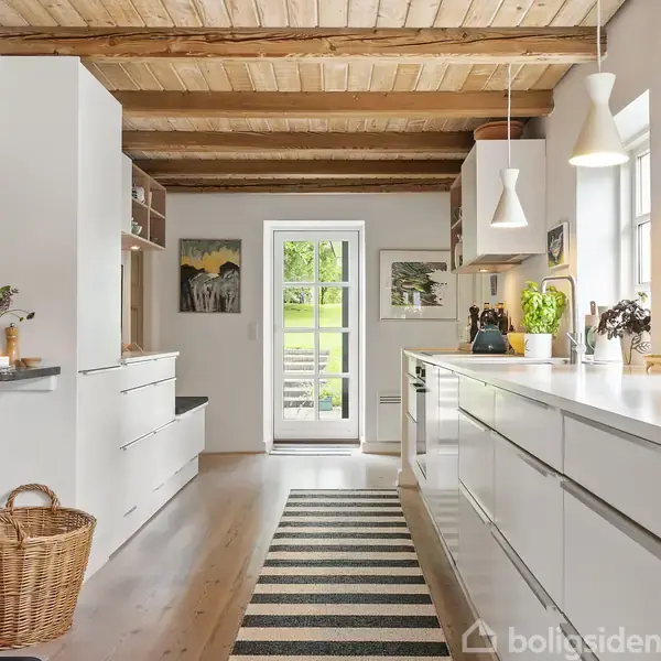 Køkken med hvide skabe og en sort-hvid stribet løber. Der er træloft og gulv. To store vinduer slipper lys ind, og en kurv står ved siden af en sort ovn.