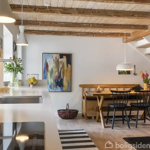 Køkken med en vask og kogeplader på en hvid bordplade. Spisebord med sorte stole og blomster i baggrunden. Træloft, trappe og kunst på væggen skaber en hyggelig atmosfære.