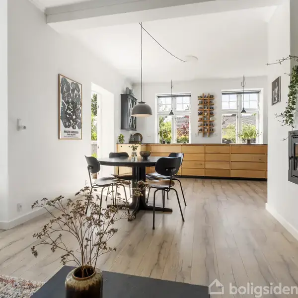 Stue med grå sofa og sofabord, udsmykket med kunst på væggen. Spisebord og stole i hjørnet. Store vinduer bringer naturligt lys ind, omgivet af grønne planter.