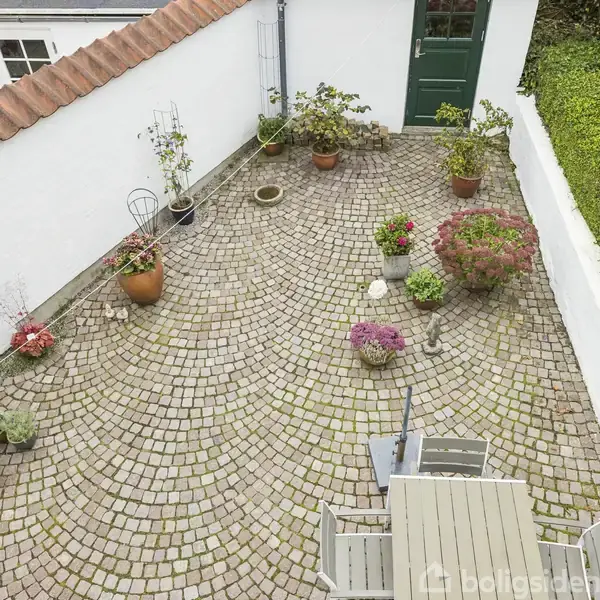 En brostensbelagt gårdhave med flere blomster i krukker og potter spredt omkring. En hvid mur til venstre og grønne hække til højre omkranser området.
