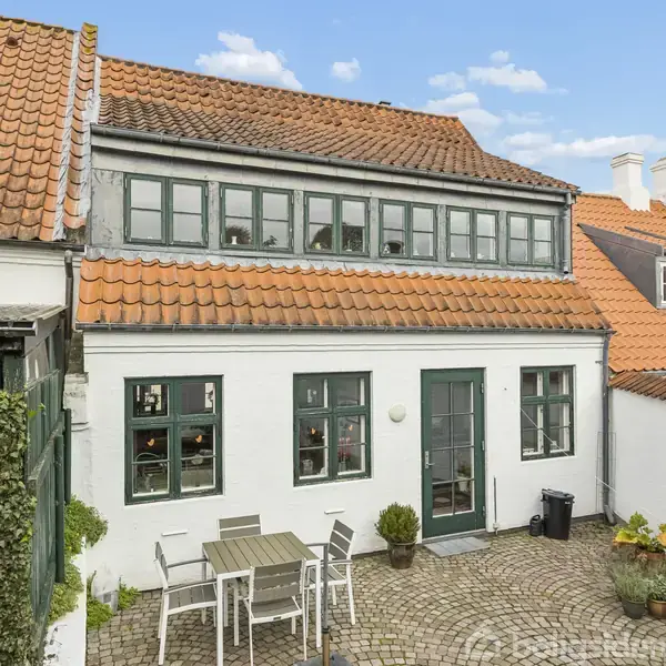 Et charmerende, hvidt hus med rødt tegltag står i en gårdhave med brosten. Et bord og fem stole er placeret foran husets grønne dør og vinduer.