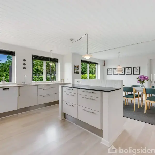 Beige køkken med en ø, hvide skabe og skuffer i moderne design. Åbent mod en spisestue med grønne stole omkring et bord, dekoreret med lilla blomster. Store vinduer giver udsigt til grønne omgivelser.